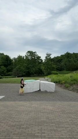The video shows a woman standing in front of two large white stone sculptures. She is wearing a yellow dress and a dark top and has a bag slung over her shoulder. She is looking at the sculptures. The sculptures are located in a gravel area with a paved walkway in the foreground. In the background, there is a grassy hill covered in trees and bushes. The sky is overcast.