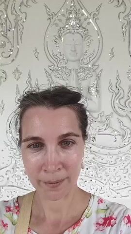 The video shows a woman in the foreground taking a selfie. She has her hair pulled back in a bun and is wearing a white shirt with a floral pattern. She is standing in front of an ornate white structure with intricate carvings and mirrored details. In the background, another woman is visible, sitting and looking down. Further in the background, trees and a cloudy sky can be seen. The overall impression is of a religious or cultural site.