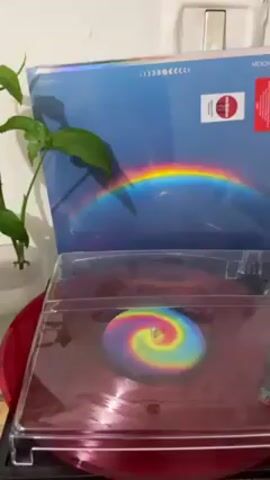 Here's a description of the video:

A vinyl record player is playing a Coldplay album called "Moon Music." The record itself is a translucent red color with a colorful spiral design in the center. The album cover is visible behind the record player. It features a blue sky with a rainbow and a small moon. There is a small plant in a white pot to the left of the record player. The scene is indoors.