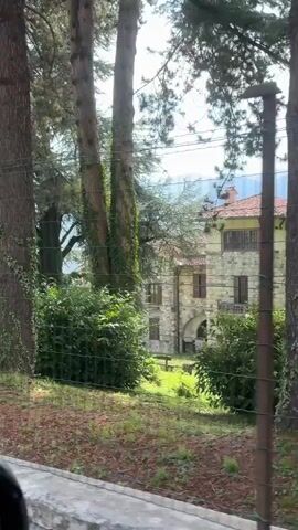 The video shows a fence with trees and foliage. A post with the number 20 on it is visible. Beyond the fence, a building and mountains can be seen. The scene appears to be outdoors in a natural setting, possibly a park or a residential area.