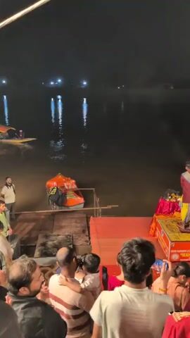 A nighttime video captures a religious ceremony on a body of water, likely a river, with the shore visible in the background. Several individuals, dressed in traditional Indian attire with maroon tops and yellow lower garments, are standing on raised platforms at the water's edge. They are performing a ritual involving fire, holding ornate lamps that emit flames and smoke. The flames are directed towards the water, creating a spectacle for the onlookers gathered on the shore. The onlookers, a diverse group of men, women, and children, are positioned in the foreground, observing the ceremony. Some are holding up their phones to record the event. The atmosphere appears festive and devotional. The water reflects the distant lights and the flames from the lamps. In the water, a small orange boat is visible, suggesting it might be used for the ceremony or for transportation. The overall scene is illuminated by the flames and some artificial lighting, creating a dramatic and spiritual ambiance.