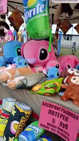 A street vendor is selling plush toys and balloons. The vendor is not visible in the video. The toys are arranged on a table and include characters like Stitch, cats, and other stuffed animals. There are also large inflatable toys shaped like soda cans and bags of chips. A sign is visible advertising a game where customers can pop balloons to win prizes. The game involves a board with balloons of different colors, and prizes are awarded based on the number of balloons popped. The environment appears to be an outdoor market or fair.
