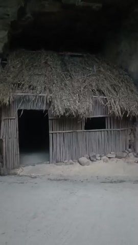 The video shows a static shot of a rustic structure resembling a hut. The hut has a thatched roof and walls constructed from vertical bamboo-like poles. There is an open doorway on the left and a small window on the right. The ground in front of the hut is sandy and a few rocks are scattered around the base. The background appears to be a dark, rocky alcove or cave. The lighting is dim. The video is 3 seconds long and appears to be a single, unedited shot.