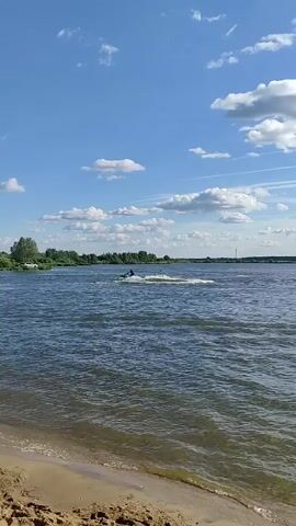 A jet ski with a person on it is seen from a sandy shore. The jet ski is moving across a body of water, leaving a wake. The sky is blue with scattered white clouds. Trees and land are visible in the distance.