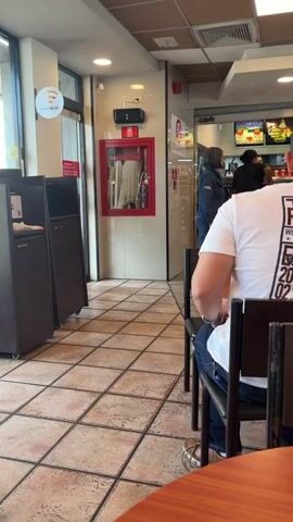 The video shows the interior of a fast-food restaurant. The camera is positioned behind a person wearing a white t-shirt, who is seated at a table. In the background, there is a counter area where employees are visible. One employee, a woman with dark hair, is seen interacting with someone behind the counter. There are menu boards displayed above the counter. The floor is tiled, and there are trash receptacles and a fire extinguisher cabinet visible along the wall. A sign indicating "Wi-Fi" is also present. The overall environment appears to be a typical casual dining establishment.