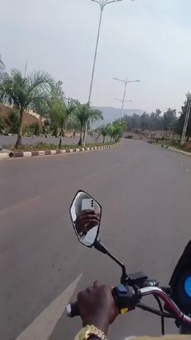 The video shows a person riding a motorcycle on a paved road. The road has a painted dividing line, and on the left are sidewalks and a line of palm trees. A white car with a yellow stripe is driving in the same direction as the motorcycle. The rearview mirror of the motorcycle reflects the rider's hand holding a phone. The rider wears a gold watch. There are hills and buildings in the distance under a cloudy sky.