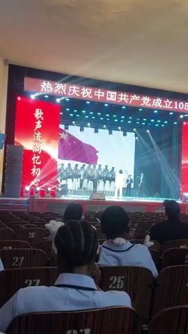 A person is singing into a microphone on a stage with a choir behind them. The stage has a large LED screen displaying a Chinese flag and then Chinese characters that appear to read "大." The choir members are dressed in black and white attire. The singer is wearing a black shirt, black pants, and a red tie. The audience is visible in the foreground.