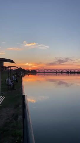 A vertical video captures a sunset over a calm body of water. The sky is filled with hues of orange, pink, and blue, with scattered clouds reflecting in the water. A metal bridge with a lattice structure spans across the water in the background. On the left bank, there is a grassy area with a few people sitting on benches under covered shelters. A dark railing runs along the edge of the embankment.