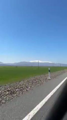 The video shows a landscape shot from a moving vehicle, likely a car, on a clear, sunny day.  The scene is dominated by a vast, flat green field stretching towards the horizon. In the distance, a mountain range capped with snow is visible under a bright blue sky.  Wooden utility poles line the roadside.  The camera appears to be positioned low within the vehicle, providing a view of the blacktop road and a strip of gravel along the edge.  The camera is moving quickly, creating a sense of speed.
