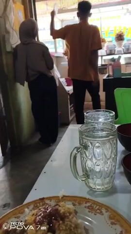 The video shows an indoor scene, likely a food stall or small eatery. In the foreground, a plate of food is visible on a white table, accompanied by two glasses of water and two dark bowls. In the background, a person wearing a headscarf and dark clothing stands near a counter, and another person in an orange shirt is working behind the counter, appearing to be in motion. The environment looks somewhat rustic, with visible wooden beams and corrugated roofing overhead. The lighting suggests it is daytime.