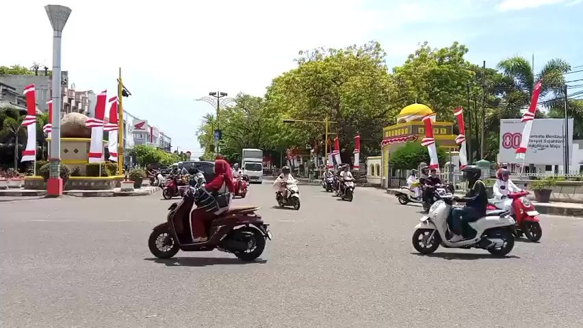 This is a daytime, medium-wide shot of a busy intersection in a town or city, with moderate traffic. Numerous motorcycles are the predominant mode of transportation, with a few cars also visible. Many of the vehicles and surrounding buildings are decorated with red and white flags, suggesting a national holiday or celebration. There are trees and streetlights lining the road. In the background, a building with a yellow dome, likely a mosque, is visible, also adorned with decorations. The sky is bright and appears clear. People on the motorcycles are diverse in age and gender, and most are wearing casual clothing. Some riders are alone, while others have passengers. The overall atmosphere appears to be one of everyday activity with an added festive element due to the decorations.