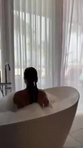 A woman is relaxing in a bathtub filled with bubbles. She is sitting facing away from the camera, with her wet hair pulled back. The bathtub is white and oval-shaped, and there is a chrome faucet next to it. Behind her, there is a large window with sheer white curtains. The overall impression is one of relaxation and luxury.