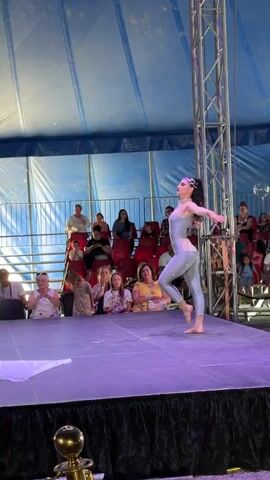 A female performer in a shimmering silver outfit with a jewel-encrusted neckline and headband is dancing on a stage. She has her hair pulled back and is barefoot. In the background, an audience is seated in tiered rows, watching the performance. A large blue and white tent structure is visible behind the audience. A piece of white fabric is draped on the stage, and what appears to be aerial silks are hanging from a rigging system. The performer moves gracefully, extending her arms and turning. The lighting on the stage is purple.