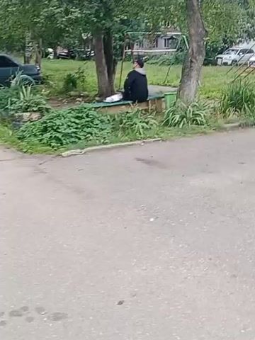 A person is sitting on a wooden bench outdoors, with greenery and trees surrounding them. The person is wearing a dark jacket with a hood up and dark pants. In front of them on the bench is a white object, possibly a bag or container. The person appears to be looking at something off-camera and then turns to smile at the camera. There is a children's swing set in the background, along with parked cars and buildings. The ground in the foreground is paved.