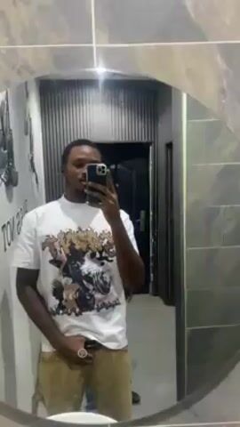 A young man is recording a selfie video in a bathroom mirror. The mirror is round and framed by gray tiles. The bathroom has gray walls with white and gray tiles. Behind the man, another young man enters the bathroom and stands behind the first man. The first man wears a white T-shirt with a large graphic on the front and khaki pants. He smiles at the camera. The second man wears a black T-shirt with a graphic on the front and a black beanie.