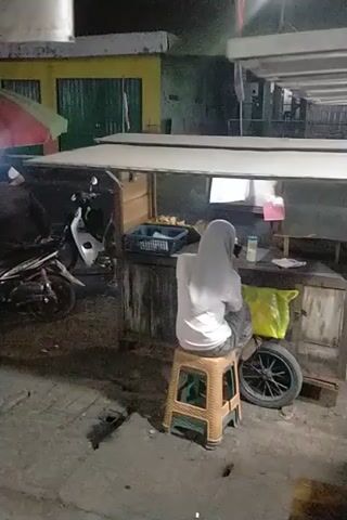 The video shows a person wearing a headscarf, likely a woman, sitting at a small outdoor stand or shop. She is facing away from the camera and appears to be engaged in some activity behind the counter. The stand is stocked with various items, including bottles, a water dispenser, and what appear to be packaged goods. A motorcycle is parked in front of the stand, and a spare tire and a yellow plastic bag are visible nearby. The environment seems to be an urban street or marketplace, with some lighting suggesting it might be evening or nighttime. The camera is shaky and the footage is of low quality, making some details difficult to discern.