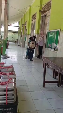 A woman in a pink hijab and white shirt, possibly a teacher, is standing in a doorway of a classroom or office. She is facing two young girls in dark green school uniforms, each carrying a backpack. The girls appear to be interacting with the woman. They are standing in a tiled hallway with lime green walls. To the left, there are stacks of metal trays with red ribbons. To the right, there is a wooden table and chairs. The hallway has windows and doors along the wall.
