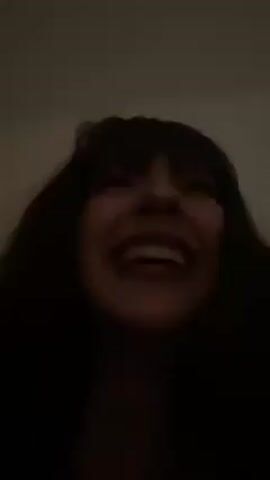 A woman with long dark hair and bangs laughs at the camera. She is wearing dark red lipstick. She covers her mouth with her hand, displaying a collection of silver rings on her fingers, some with colorful stones. The background is a light beige or white wall. The lighting is dim.