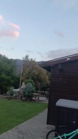 The video shows a lawn at dusk. There is green grass, trees, a trimmed hedge, and a palm tree. Mountains are visible in the background. The sky has pale pink and gray clouds. The video pans across the scene. The edges of a building or patio are visible in the foreground.