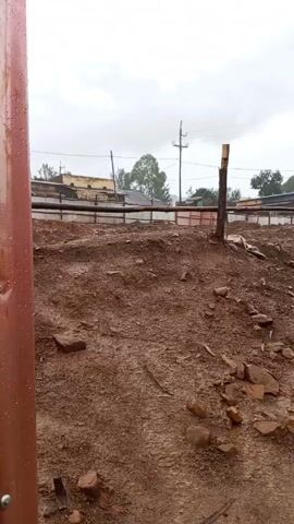 The video shows a patch of muddy land on a rainy day. The ground is covered in rocks and debris. In the background, there are some buildings and trees. A utility pole stands in the distance. A red corrugated metal structure is partially visible on the left side of the frame.