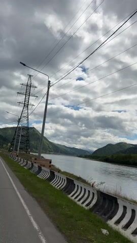 The video shows a landscape on an overcast day. A gray highway stretches into the distance. Beside the highway, a black and white striped guardrail separates the road from a grassy area and a wide river. In the background, rolling green hills line the river. A tall, black power line tower is situated in the grassy area between the highway and the river. A street lamp is mounted on a gray pole beside the power tower. The sky is filled with gray and white clouds with patches of blue.
