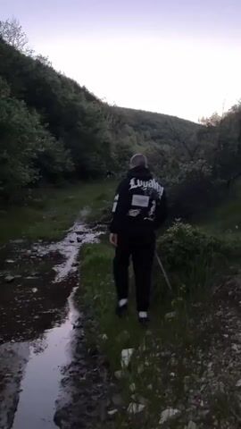 Two men are walking in a grassy field between two wooded hills. The man at the front of the frame wears a black hoodie and black pants. The second man is slightly obscured by the first but appears to be carrying a light-colored object, perhaps a skateboard. The sky above them is a gradient of blue and pink, suggesting early morning or late evening.  The environment is dark with the sun on the horizon.