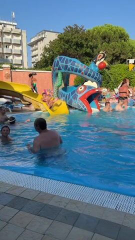 A video shows a pool with several people enjoying the water. In the background, there is a blue and red serpent-shaped water slide with a yellow slide extending from its side. A young boy slides down the yellow slide and splashes into the pool. Other individuals are swimming, wading, and interacting in the water. The environment appears to be an outdoor swimming pool area with trees, buildings, and a clear blue sky visible in the background.