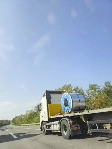 The video shows a highway scene. In the first frame, a white truck with a yellow load is driving on the road. The second frame shows the truck from a slightly different angle. The sky is clear and blue. The road is an asphalt highway with white lane markings. There are trees and foliage on the side of the road. The video appears to be taken from a moving vehicle.