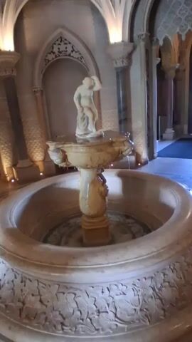 A statue of a person stands atop a stone fountain. The fountain is in the center of a room with arched ceilings and ornate columns. The fountain is lit by spotlights from above.