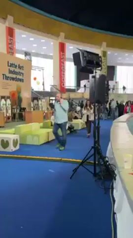 The video shows the interior of a large circular building, possibly a convention center or exhibition hall, with multiple levels and a high dome-shaped ceiling. The ground floor is filled with various booths and displays, with people walking around and interacting with the exhibits. On the mezzanine level, people can be seen walking along the railing, observing the activity below. The building's architecture features columns and arched windows, creating a sense of openness. Banners with text are hung from the mezzanine level. A sound system is visible near the start of the video. The overall impression is a bustling and crowded environment, with a wide range of activities taking place in a large, well-lit space.