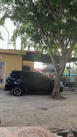This video captures an outdoor scene with a black minivan parked under a large tree. The minivan is the central object, positioned on a dirt and gravel ground. To the left of the minivan is a yellow building with glass doors and windows, and signage that partially reads "BSI." Behind the minivan, a red-clad figure can be seen under a covered area, and a bicycle is parked nearby. The scene is shaded by the dense foliage of the tree, with sunlight filtering through the leaves, suggesting it is daytime, possibly late afternoon. The overall setting appears to be a relaxed, possibly semi-rural or commercial area, given the building and the presence of vehicles. The ground is uneven and strewn with small stones and debris. The lower portion of the frame shows a tiled surface, possibly a patio or walkway. The weather appears to be clear, with no visible rain or overcast skies. The location context indicates Lampeuneurutblang, Indonesia.