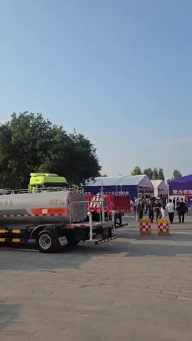 The video shows multiple large trucks parked on a paved surface. Some of these trucks appear to be tanker trucks, with large cylindrical tanks mounted on their chassis. Some of the tanker trucks are white, while others are gray and orange. At least two of the trucks have cranes mounted on them. One crane is red and yellow, and another is yellow. There are flags on tall poles in the background, as well as trees and a bright sky with the sun visible. One truck has a red spherical decoration on top of its crane. The scene appears to be outdoors, possibly at an event or exposition, given the presence of tents in the background. There are a few people visible, walking around the trucks and the area.