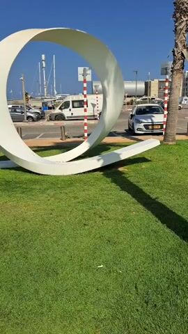 A white, modern sculpture resembling a rounded "Q" is situated on a patch of green grass under a clear blue sky. To the left of the sculpture, a street is visible with several vehicles, including white vans and cars, and a small bicycle. In the background, there are buildings and the masts of sailboats, suggesting a location near a harbor or marina. A palm tree stands to the right of the sculpture, casting a shadow.