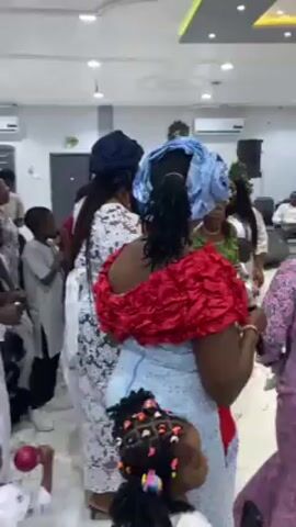 The video shows a gathering of people in a bright, well-lit room, likely a reception hall, with white walls and modern lighting fixtures. The group consists of both adults and children, dressed in a variety of festive attire, including traditional clothing with lace and ornate head coverings. There is a mix of colors and patterns in their outfits, such as whites, blues, reds, greens, and pinks. Many individuals appear to be engaged in conversation and movement, suggesting a celebratory atmosphere, possibly a party or event. The scene seems lively and animated.