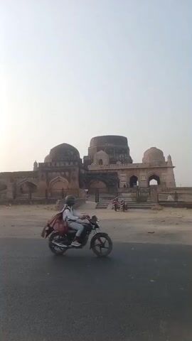 The video shows a complex of weathered, stone structures with domed roofs, situated near a paved road. The structures are enclosed by a metal fence and appear to be historical buildings. Two motorcycles are parked near the entrance, and a person is visible through one of the archways. The video pans to the right, showing the length of the structures and a tree in the foreground with the sun shining through its branches. Another smaller tree is further down the road. A motorcycle travels from left to right on the road in the direction of the structures. The sky is overcast, creating a muted, dusty atmosphere. The road is visible in the foreground, leading towards the historical site.