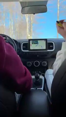 The video shows the interior of a moving Hyundai Venue car, with a view through the windshield. Two people are visible: the driver wearing a maroon sweatshirt and a passenger in a white sweatshirt. The driver's hands are on the steering wheel, while the passenger holds a yellow object and a phone. The car's navigation screen is displaying a route. Outside the car, there's a snowy landscape with trees and a blue sky. The car seems to be driving on a road covered in snow, past snow-covered buildings.