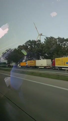 The video shows a view from inside a vehicle, looking out the window onto a highway. The sky is overcast and appears to be either early morning or late evening. On the highway, there are several large trucks. In the foreground, a reflection shows a person sitting in the vehicle, holding a white object. The person is wearing a blue shirt. The trucks on the highway are a mix of yellow and white trailers. One truck is carrying a very large, white, curved object, possibly a component of a wind turbine or similar industrial equipment. The road is paved and appears to be a multi-lane highway. There are trees and vegetation visible alongside the road. A sign with blue and white symbols is visible on the right side of the road, indicating a rest stop or service area. The video captures movement as the vehicle travels along the highway.