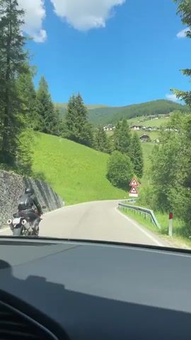 A motorcycle is seen from a car in a scenic mountainous region with green hills and trees under a blue sky with clouds. The motorcycle is black and ridden by a person wearing a black outfit. There are road signs on the right side of the road, including a warning sign with a zigzag arrow and a sign indicating a sharp curve. The road is paved and curves to the right. The car is moving and filming the motorcycle.