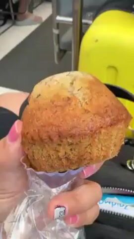 A person is holding a banana nut muffin in a clear plastic wrapper. The muffin is large and golden brown with visible nuts. The person has pink nail polish with a design on one nail. The background includes a yellow suitcase, a metal bar, and a glimpse of someone wearing shoes. The scene appears to be inside a vehicle or transportation setting due to the yellow luggage and metal bar. There is some noise, possibly from the vehicle's engine.