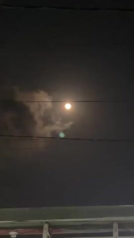 The video shows a nighttime sky with a full moon partially obscured by clouds. Two power lines run horizontally across the frame. A blurry, greenish light hovers between the power lines, closer to the viewer than the moon. The bottom of the frame shows the top of a structure, possibly a building or vehicle, with some metal railings. The overall impression is of an outdoor scene at night, with celestial bodies and man-made infrastructure visible.