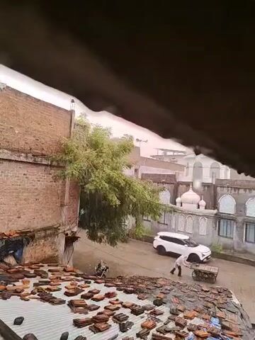 The video shows a street scene during what appears to be a rainstorm. In the foreground, a rooftop is covered with many brown roof tiles, some of which are scattered. A large green tree with dense foliage is visible on the left side of the frame, leaning slightly into the street. Behind the tree, a brick building is present. Across the street, a white SUV is parked. A person dressed in white is pushing a cart. In the background, there are several buildings, including one with a dome, suggesting a residential or potentially religious area. Raindrops are visible, indicating the ongoing precipitation.