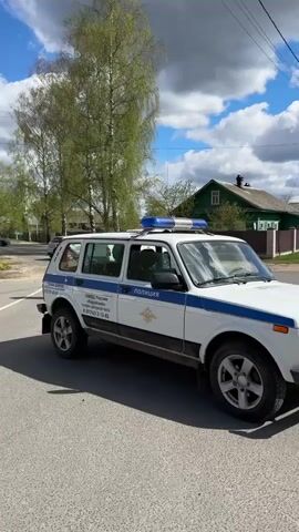 The video shows a police checkpoint on a road. A white Lada Niva police vehicle with blue stripes and a light bar is parked on the right side of the road. Several police officers in dark uniforms are standing near a white pickup truck that has been pulled over. Pedestrians walk on the sidewalk along the road. The sky is partly cloudy with patches of blue. Trees line the street. A "No Stopping" sign is visible on a utility pole. Other cars drive past the checkpoint.