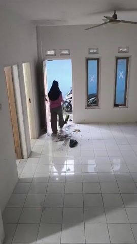 A person wearing a hijab and a pink shirt is sweeping a tiled floor in front of an open doorway. Debris is scattered on the floor near the doorway. The room has white walls and a ceiling fan. There are two windows with blue coverings on the right wall.