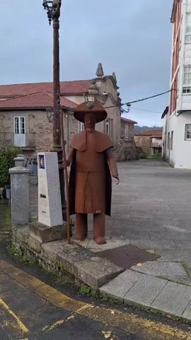 The video shows a statue on a stone platform in a town square. The statue is made of rusted metal and depicts a pilgrim with a staff, wide-brimmed hat, and cape.  Next to the statue is a stone marker with a yellow scallop shell symbol, likely indicating the Camino de Santiago pilgrimage route.  Behind the statue is a stone building with a red tile roof. To the right is a taller building with a white facade and red brick accents. The square is paved with gray stones. There are ornate street lamps. The sky is overcast.
