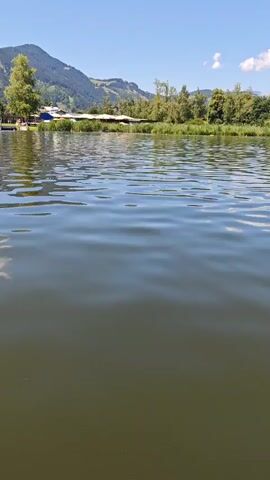 The video shows a body of water with a shoreline visible in the background. On the shore, there are several pedal boats docked. In the foreground, part of a boat is visible, with two people sitting in it, facing away from the camera. The scene appears to be a recreational area. The water is calm with ripples on the surface. Trees and hills form the landscape behind the shoreline. The sky is clear and blue.