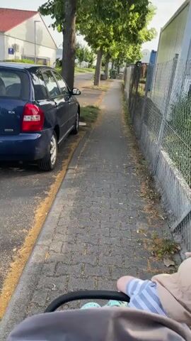 This video shows a first-person perspective of someone pushing a baby stroller along a paved sidewalk. The stroller contains a baby wearing a light-colored hat with ear-like embellishments and a striped shirt. The stroller itself is gray with a green woven basket underneath.

The setting appears to be a suburban or light industrial area in Germany, possibly Nidda. A dark blue car is parked on the left side of the street, adjacent to the sidewalk. The sidewalk is bordered by a chain-link fence on the right, behind which is some greenery. Mature trees with full, green canopies line the sidewalk, casting dappled shade.

In the background, across the street, there are buildings, including one with a red tiled roof and what appears to be a parking sign. Further down the street, there are more trees and some industrial-looking containers. The lighting suggests it is daytime, likely late afternoon or early evening, given the warm light. The overall mood seems peaceful and routine, likely a leisurely walk. There is no visible text within the video frame.