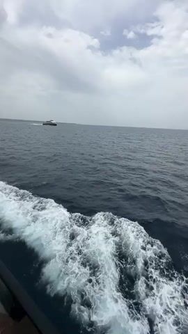 The video shows a view from a moving boat on the ocean. In the foreground, white frothy waves are visible on the surface of the dark blue water, indicating the boat's wake. In the middle ground, a white passenger ferry is seen moving across the water in the distance. The sky above is overcast with gray clouds.