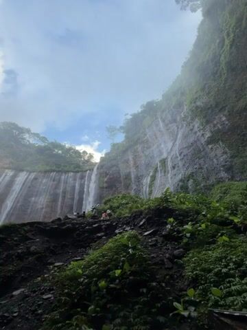 The video shows a majestic waterfall cascading down a steep, moss-covered cliff. The water falls in numerous thin streams, creating a curtain of white against the dark rock face. Lush green vegetation covers the slopes surrounding the waterfall, with trees visible at the top of the cliff. The sky is partly cloudy, with patches of blue peeking through the white clouds. In the foreground, a person stands on a rocky outcrop, with their arms raised in a gesture of exhilaration. They are silhouetted against the backdrop of the waterfall and the sky. Other people can be seen further back on the rocky path, also appearing to be admiring the waterfall. The overall impression is one of natural beauty and awe.