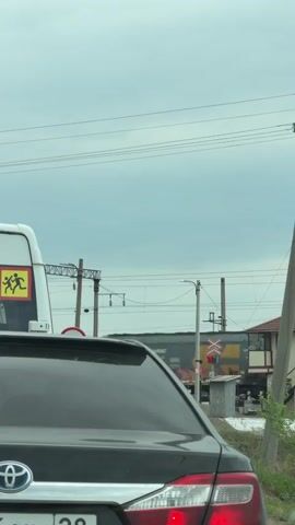 From the perspective of someone inside a dark car with a Toyota emblem on its trunk, the video captures a view of a level crossing with a slow-moving freight train passing. The railway signal lights are red. To the left, a bus or large van with a yellow and black sign depicting running children is visible. Overhead are power lines against a cloudy sky. On the right, there's a small building alongside the tracks. The camera remains still throughout the short clip.