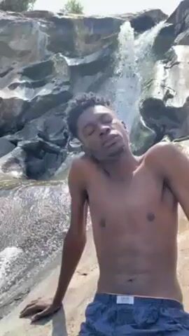 A shirtless young Black man with short curly hair poses while standing near a rocky waterfall in what appears to be a natural outdoor setting. He is wearing dark patterned shorts. The backdrop features a small waterfall cascading over large, gray rocks, with vegetation visible at the top. The weather seems sunny and bright. The man is the central focus of the shot, looking at the camera with a relaxed expression.